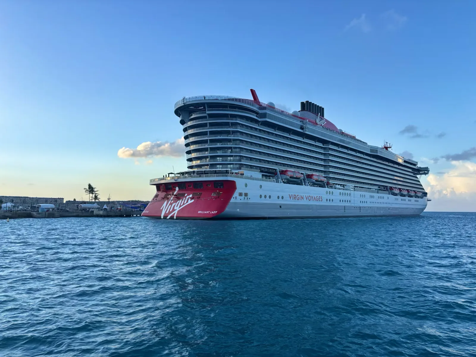 A cruise ship docked at a port pier under a clear sky, calm water in the foreground