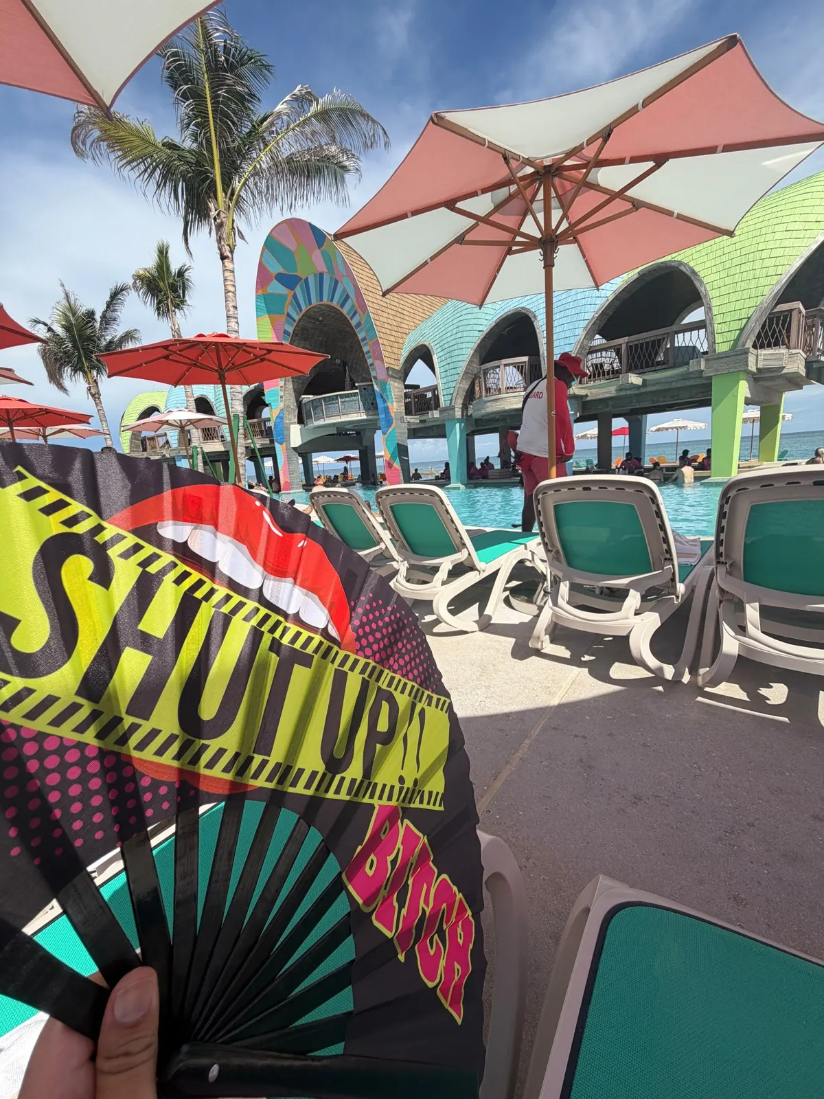 A cheeky Shut Up Bitch hand fan held up in front of the Royal Beach Club pool