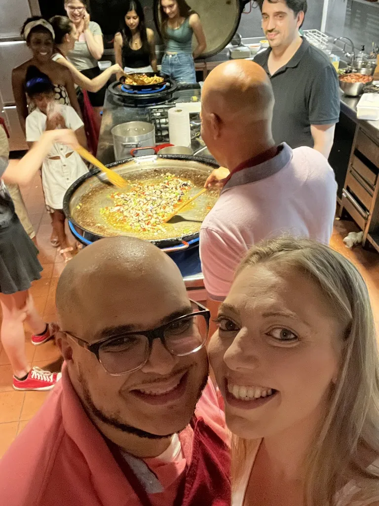 Miguel and Ashley taking a selfie at a paella cooking class while the chef stirs a large pan